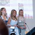 Drei Personen in traditioneller Tracht stehen mit Mikrofonen im Vordergrund und konzentrieren sich auf das Hauptprodukt oder die Idee, um die es gerade geht.