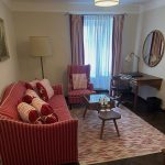Wohnzimmer mit Sofa und Sessel, rundem Spiegel, Schreibtisch mit Lampe, Couchtisch, Teppich, Stehlampe und Fenster mit Vorhängen.