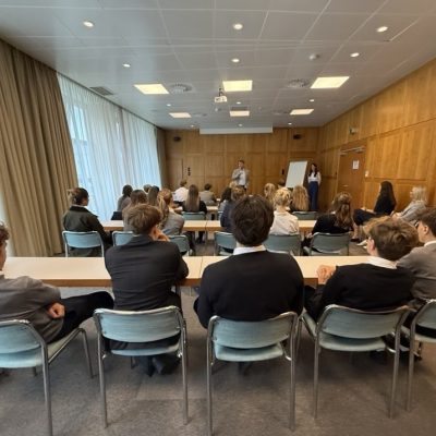 Schüler in Schuluniformen sitzen an ihren Schreibtischen und blicken zu zwei Referenten in der Nähe einer Tafel.