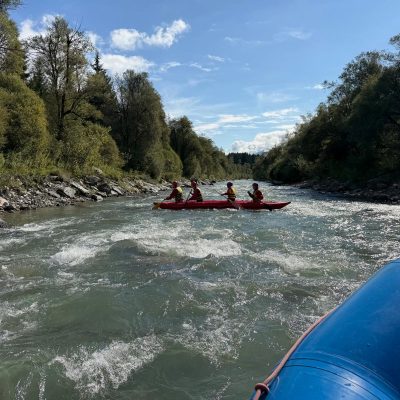 Menschen, die Helme und Schwimmwesten tragen, paddeln auf einem Floß, im Vordergrund ist ein weiteres Floß zu sehen.