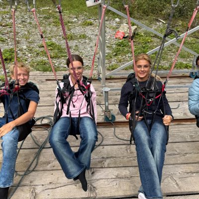 Vier Personen sitzen in Gurt-Sitzen auf einer Holzplattform und bereiten sich auf eine Aktivität im Freien vor.