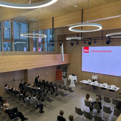 Die Menschen sitzen vor einer großen Leinwand, auf der das Wort „Urlaubsanspruch“ zu sehen ist; eine Person sitzt vorne an einem Tisch.