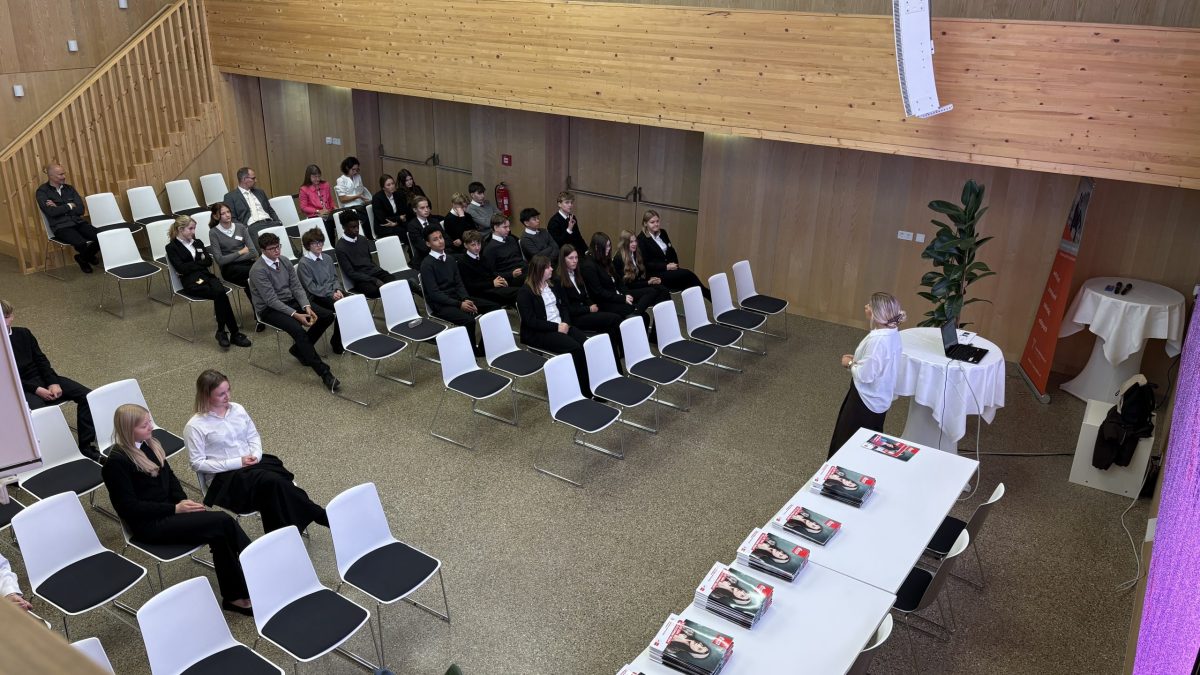Jemand hält vor einer Gruppe einen Vortrag, während auf den Tischen ordentlich gestapelte Bücher ausliegen.