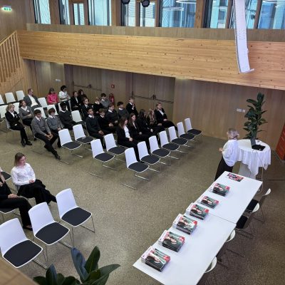 Jemand hält vor einer Gruppe einen Vortrag, während auf den Tischen ordentlich gestapelte Bücher ausliegen.