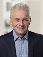 Eine Person, die eine Anzugsjacke und ein Hemd mit Knopfleiste trägt und lächelt.