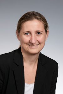 Person, die einen Blazer und ein Oberteil trägt und in die Kamera lächelt.