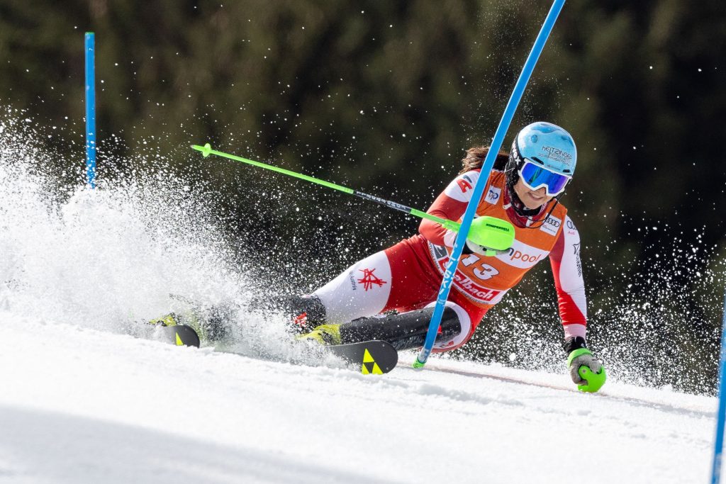 Ein Skifahrer in rot-weißer Ausrüstung fährt eine scharfe Kurve um ein blaues Slalomtor auf einem verschneiten Hang. Schnee spritzt beim Manövrieren auf. Der Skifahrer trägt Helm, Schutzbrille und hält neongrüne Skistöcke in der Hand. Im unscharfen Hintergrund sind Bäume zu sehen. Ein Skifahrer in rot-weißer Ausrüstung fährt eine scharfe Kurve um ein blaues Slalomtor auf einem verschneiten Hang. Schnee spritzt beim Manövrieren auf. Der Skifahrer trägt Helm, Schutzbrille und hält neongrüne Skistöcke in der Hand. Im unscharfen Hintergrund sind Bäume zu sehen.