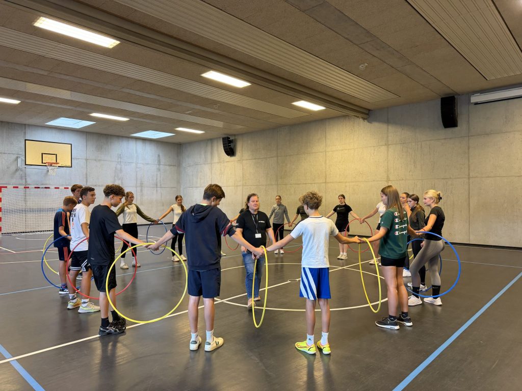 Die Teilnehmer stehen im Kreis und halten sich im Rahmen einer Teambildungsmaßnahme durch Hula-Hoop-Reifen hindurch an den Händen. Die Teilnehmer stehen im Kreis und halten sich im Rahmen einer Teambildungsmaßnahme durch Hula-Hoop-Reifen hindurch an den Händen.