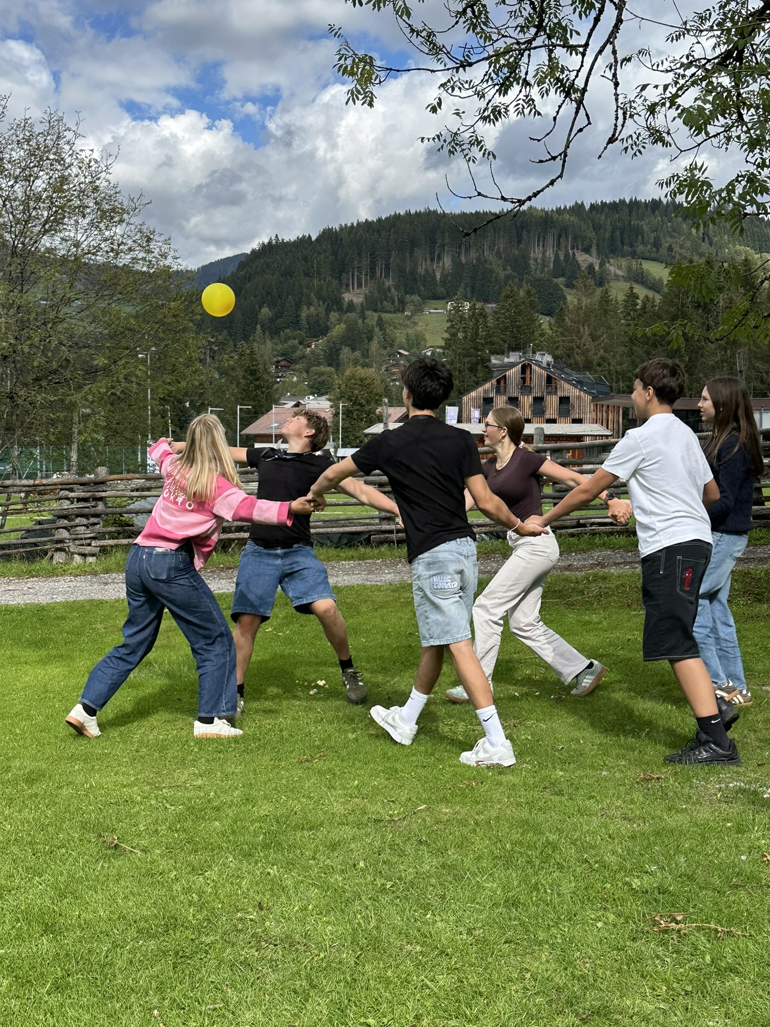 Sechs Jugendliche spielen Ball im Kreis, im Hintergrund bewaldete Berge und ein Gebäude.