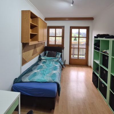 Ein ordentliches Schlafzimmer mit einem Einzelbett auf der linken Seite, mit blauer Bettwäsche. Über dem Bett befinden sich Holzregale. Ein Fenster und eine Glastür öffnen sich zu einem kleinen Balkon mit Aussicht. Auf der rechten Seite gibt es ein grünes Regal und einen weißen Schreibtisch.
