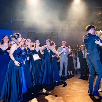 Beim Laurenz-Steiner-Gedächtnislauf 2024 tanzt eine Gruppe in Abendgarderobe bei einer Indoor-Veranstaltung. Mehrere Frauen in langen dunklen Kleidern stehen Schlange und schauen zu, während zwei Paare in der Nähe vorbeigleiten. Die Kulisse ist schwach beleuchtet und die rauchige Atmosphäre verleiht der eleganten Veranstaltung einen Hauch von Geheimnis.