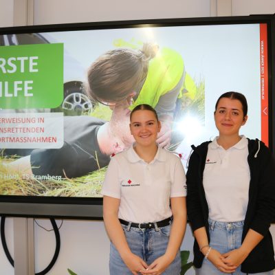 Zwei junge Frauen in weißen Hemden stehen lächelnd vor einem Bildschirm, auf dem während der Infotage eine Erste-Hilfe-Präsentation mit dem Titel „ERSTE HILFE“ läuft. Das Bild auf dem Bildschirm zeigt eine am Boden liegende Person, der geholfen wird, im Hintergrund sieht man Grün.