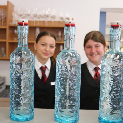 Während der Tage der offenen Tür stehen zwei Personen in Uniform hinter einer Reihe dekorativer, blauer Wasserflaschen mit floralem Muster auf der Theke. Im Hintergrund sind Regale mit Gläsern zu sehen, die zur einladenden Atmosphäre beitragen.