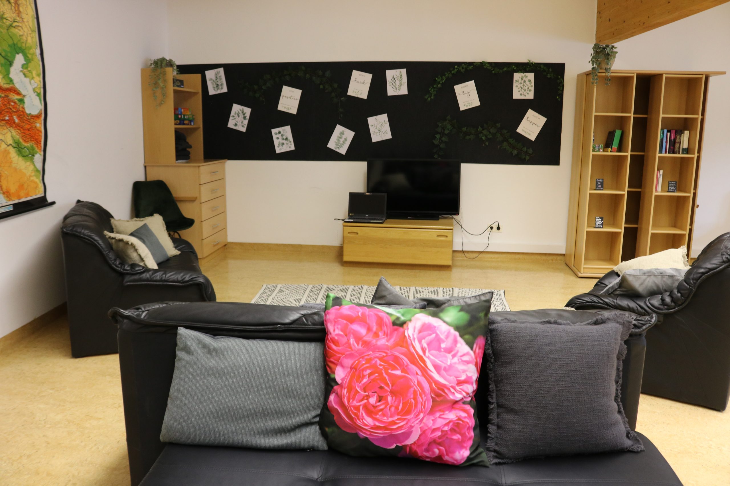 Ein Wohnzimmer mit einem schwarzen Sofa und rosa Kissen mit Blumenmotiven. Es gibt zwei weitere Sofas, eine Tafel mit angepinnten Blättern, einen Fernseher auf einem Holzständer und Bücherregale mit Pflanzen. Der Boden ist aus Holz und in der Mitte liegt ein Teppich.