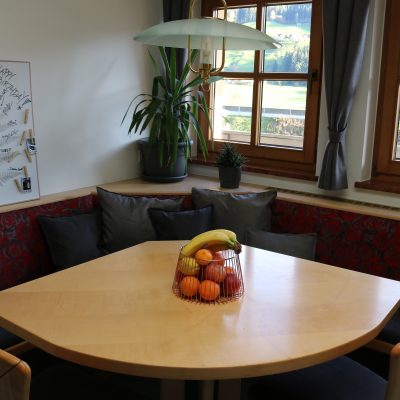 Eine gemütliche Küchenecke verfügt über einen hellen Holztisch mit einem Obstkorb voller Bananen und Orangen. Zu den Sitzgelegenheiten gehört eine rot gemusterte Bank mit schwarzen Kissen. In der Ecke steht eine Pflanze und die Fenster geben den Blick auf eine grüne Landschaft frei.