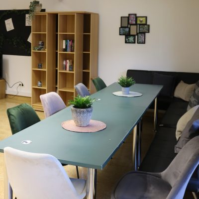 Ein Konferenzraum mit einem grünen Tisch und mehreren Stühlen. Auf dem Tisch stehen Topfpflanzen und Tischsets. An der Wand steht ein Bücherregal mit Büchern und Deko, in der Ecke steht ein Sofa. Zur Wanddekoration gehört eine Collage aus gerahmten Bildern.