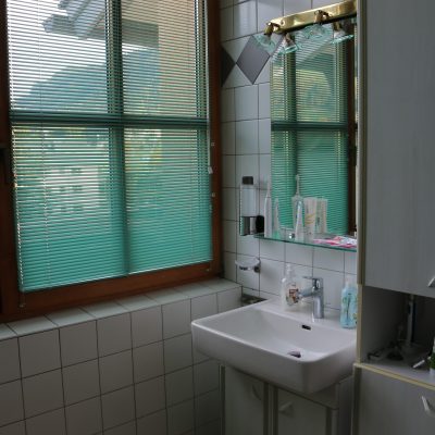 Ein Badezimmer mit einem großen Fenster und geschlossenen Jalousien, einem Waschbecken und einem Schrank. Toilettenartikel sind ordentlich auf dem Waschbecken angeordnet. Die Wände sind gefliest und über dem Waschbecken befindet sich ein Spiegel mit Beleuchtung. Der Blick nach draußen ist teilweise sichtbar.