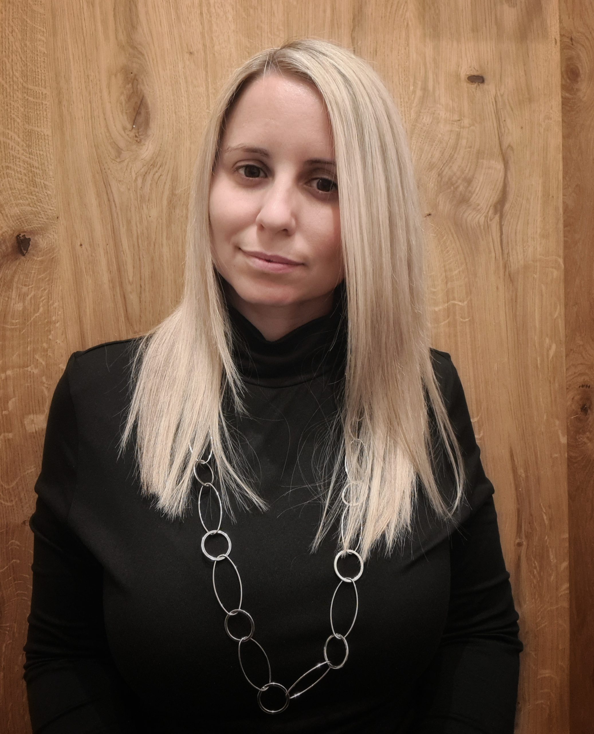 Eine Person mit langen blonden Haaren posiert vor einem hölzernen Hintergrund. Sie trägt einen schwarzen Rollkragenpullover und eine lange Halskette mit runden Gliedern.