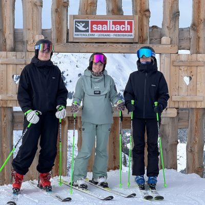 Drei Personen in Skiausrüstung posieren mit Skiern und Stöcken in der Nähe eines "Saalbach"-Schildes, das auf die Ski WM 2025 hinweist.