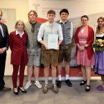 Gruppe posiert; drei in traditioneller bayerischer Kleidung, einer zeigt eine Diplomarbeit oder eine Matura-Präsentationsurkunde, ein anderer hält Blumen.