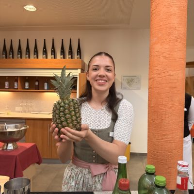 Person, die eine Ananas mit Flaschen und Bargeschirr hält, was auf eine Feier der Vorprüfungen 2025 hindeutet.