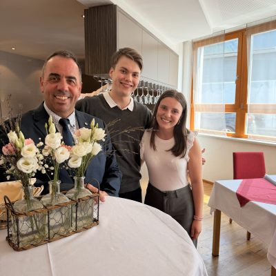Menschen, die ihren Erfolg bei Vorprüfungen von Reife- und Diplomprüfungen 2025 feiern, stehen an einem Tisch mit Blumen.
