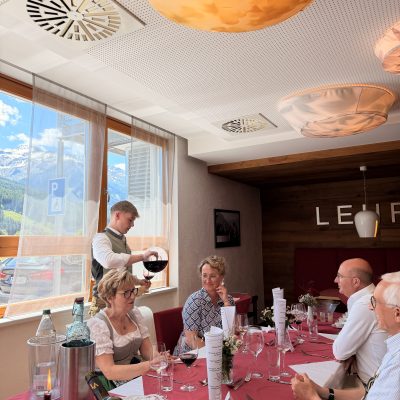 Ein Kellner schenkt Wein an einem Restauranttisch ein, während die Leute über Vorprüfungen, Reife- und Diplomprüfungen 2025 diskutieren.
