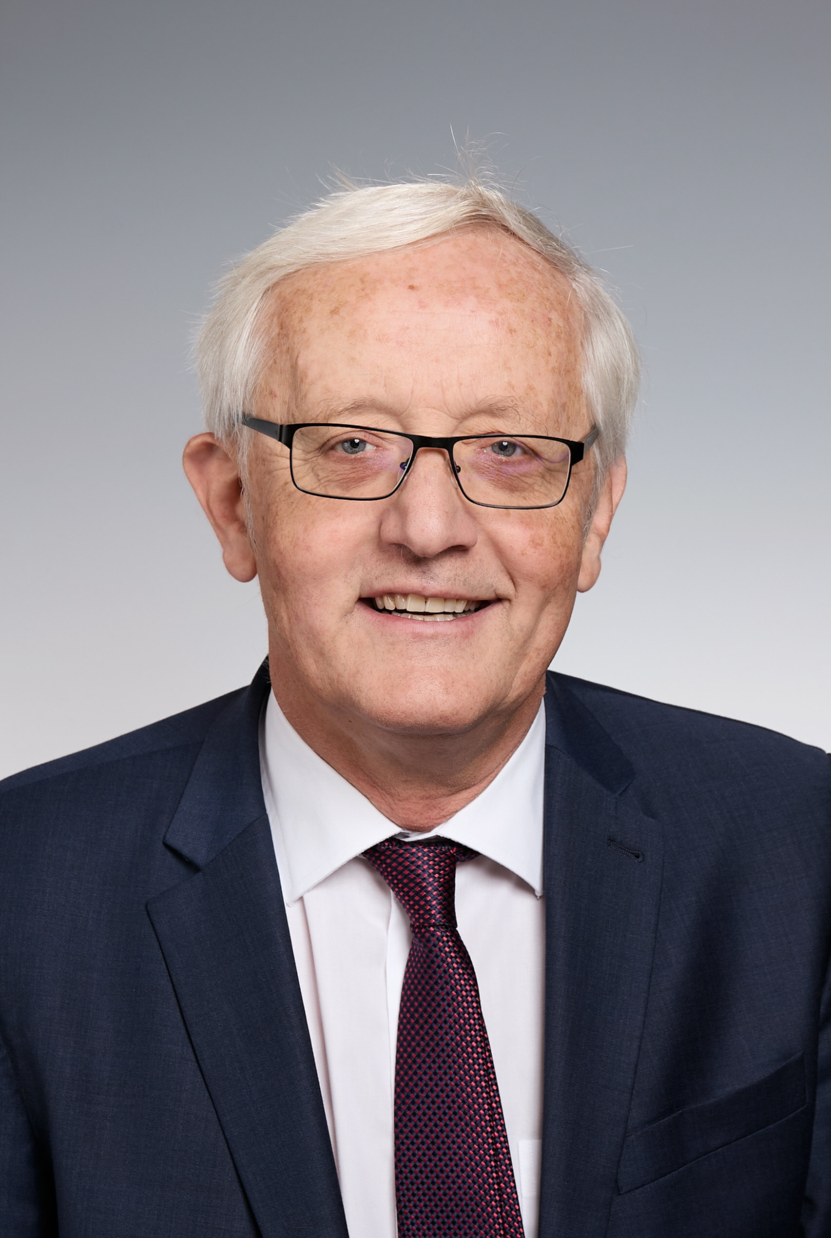 An older man with white hair and glasses is wearing a navy suit, white shirt, and red patterned tie. He is smiling slightly against a gray background.
